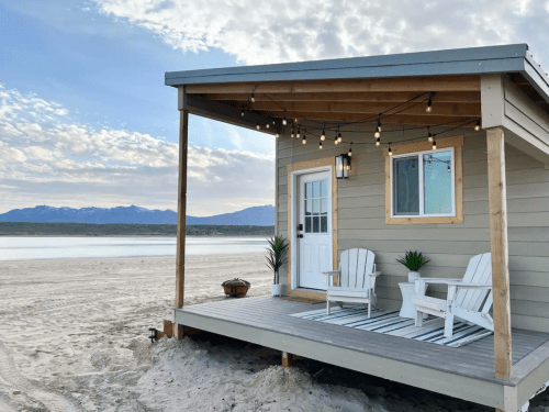 A cozy beach house with a porch, two chairs, and string lights, set against a scenic lake and mountain backdrop.